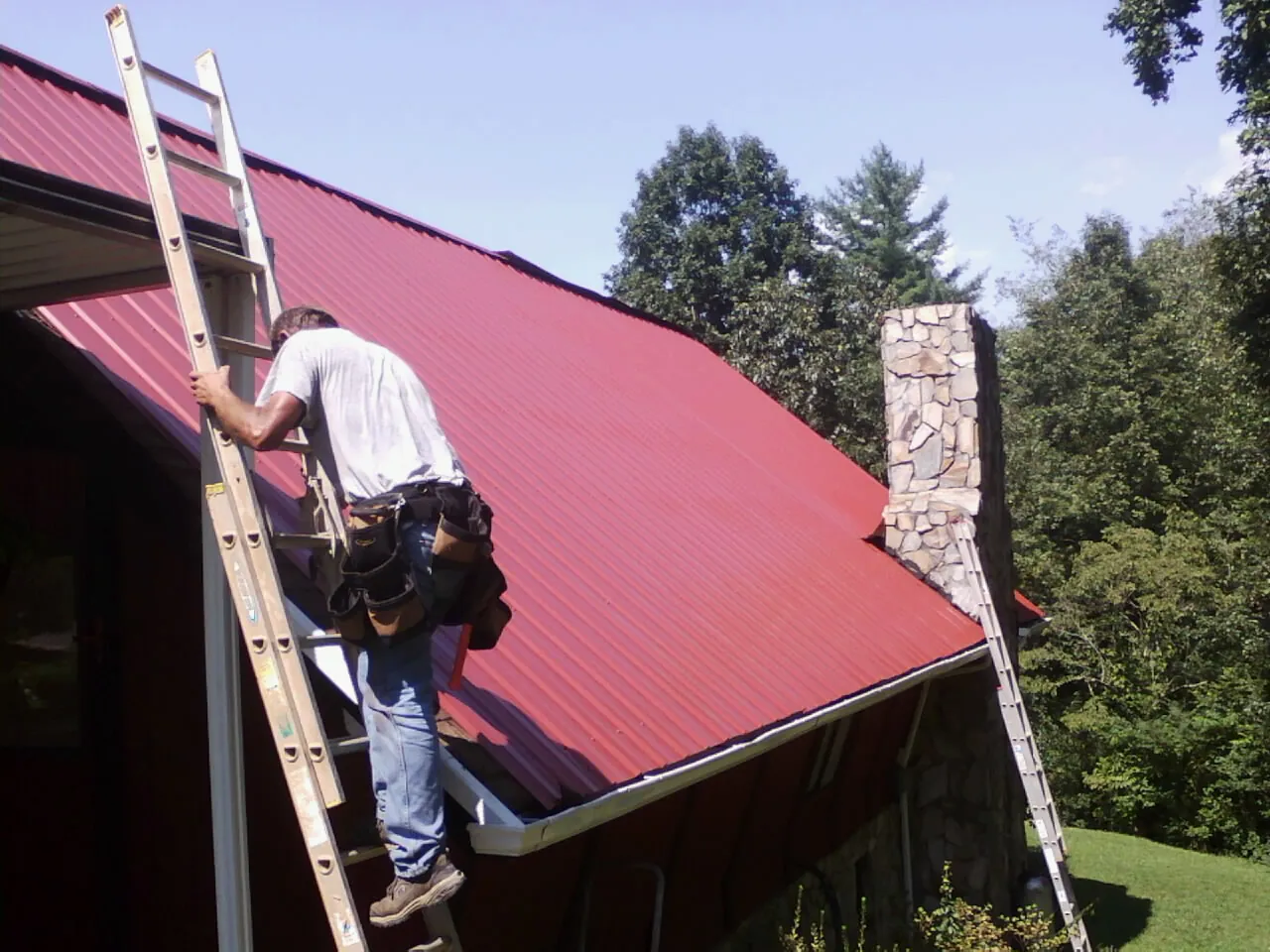 Roofer on a ladder beside a red metal roof and stone chimney — Soffit & Fascia Repair by Roofing Brothers in La Follette